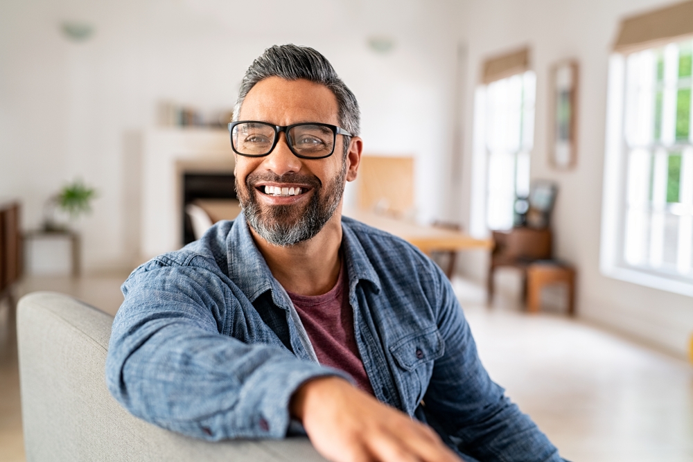 A smiling man with a beard and glasses sits on a couch, representing tooth replacement in Castle Rock at Castle Rock Family Dental